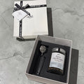 A gift set containing a jar of 'flora daydream' herbal tea and a black-handled spoon, presented in an open white box next to its closed lid on a gray concrete surface.