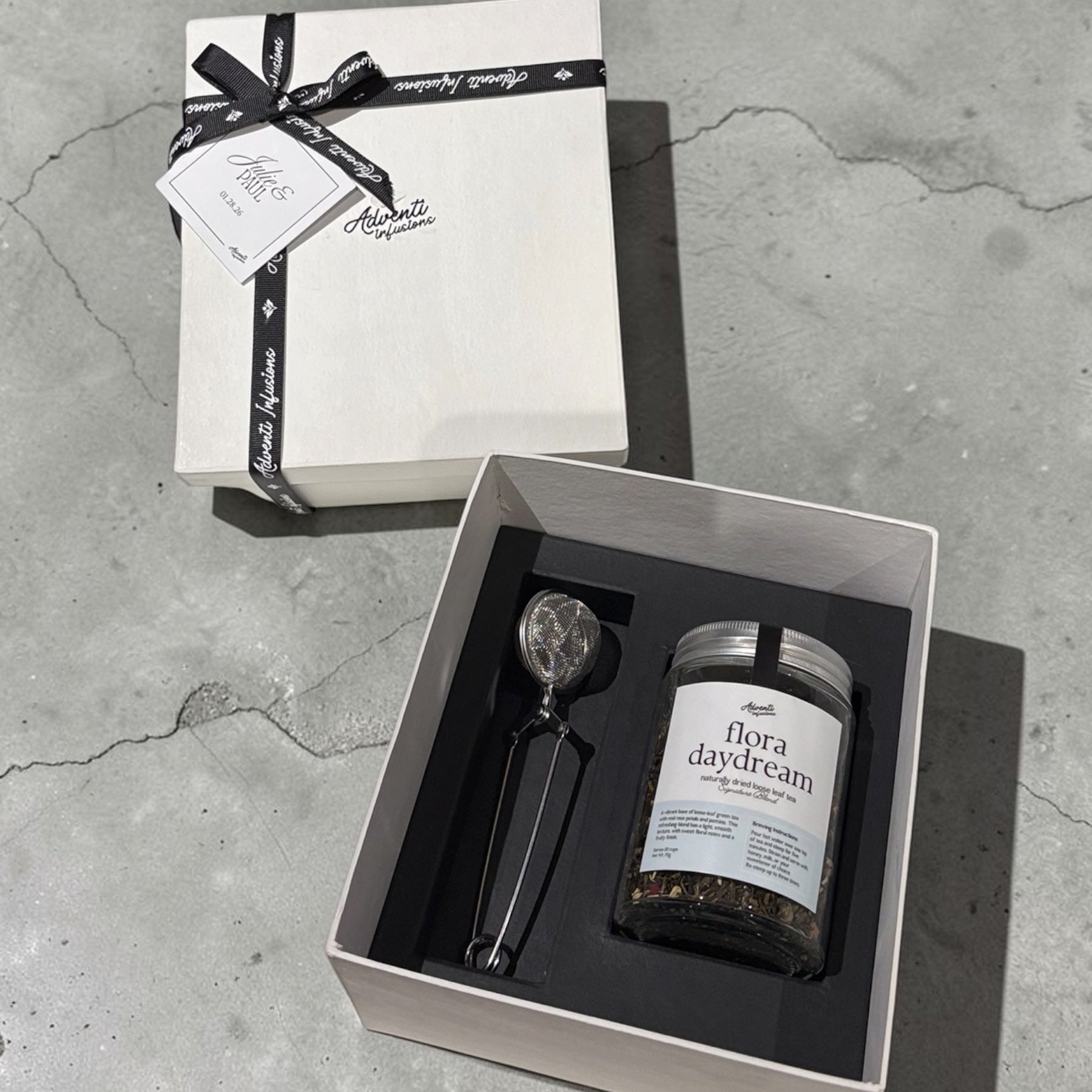 A gift set containing a jar of 'flora daydream' herbal tea and a black-handled spoon, presented in an open white box next to its closed lid on a gray concrete surface.